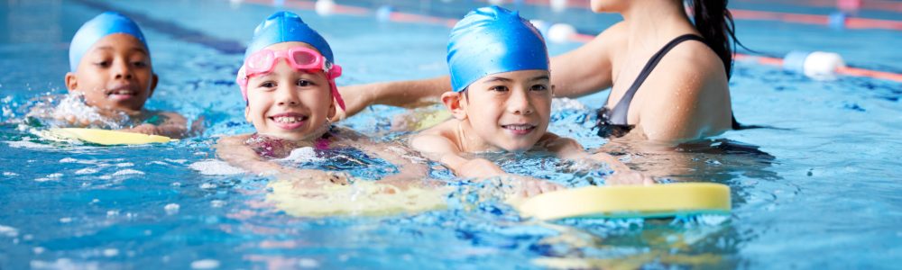 Female Coach In Water Giving Group Of Children Swimming Lesson In Indoor Pool